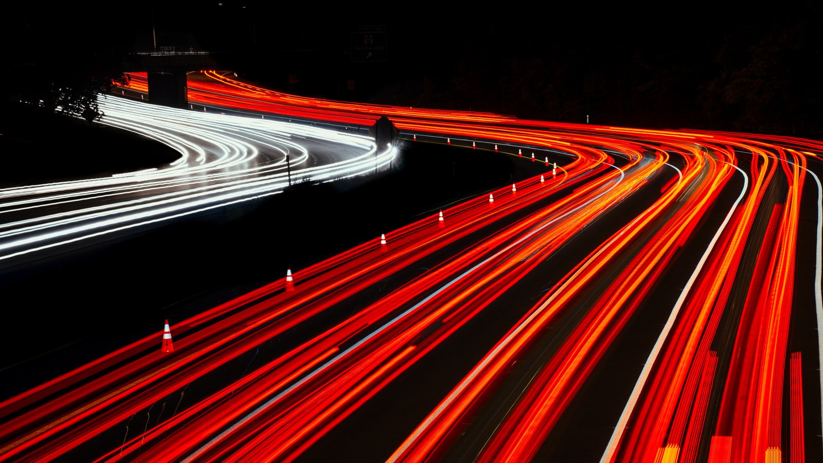A long exposure looking down on a multilane divided highway at night, every lane filled with dense red tail light trails and a line of white headlight trails curving away on the far carriageway, with a row of small traffic cones marking a taper across the near lanes. No person, registration plate, legible sign, agency marking or identifiable place appears anywhere in the frame. A generic view of nighttime highway traffic and coned lanes, not a photograph of this event.