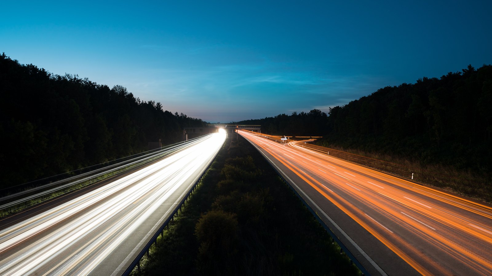 A long exposure of a divided highway at dusk seen from above, the near carriageway carrying white headlight trails and the far carriageway carrying orange tail light trails, both running freely between a scrubby median with a steel guardrail and dark wooded verges under a deep blue sky, with a low overpass crossing in the far distance. No person, vehicle, registration plate, legible sign, lettering, agency marking or identifiable place appears anywhere in the frame. A generic illustration of an open highway carrying traffic in both directions, not a photograph of this event.
