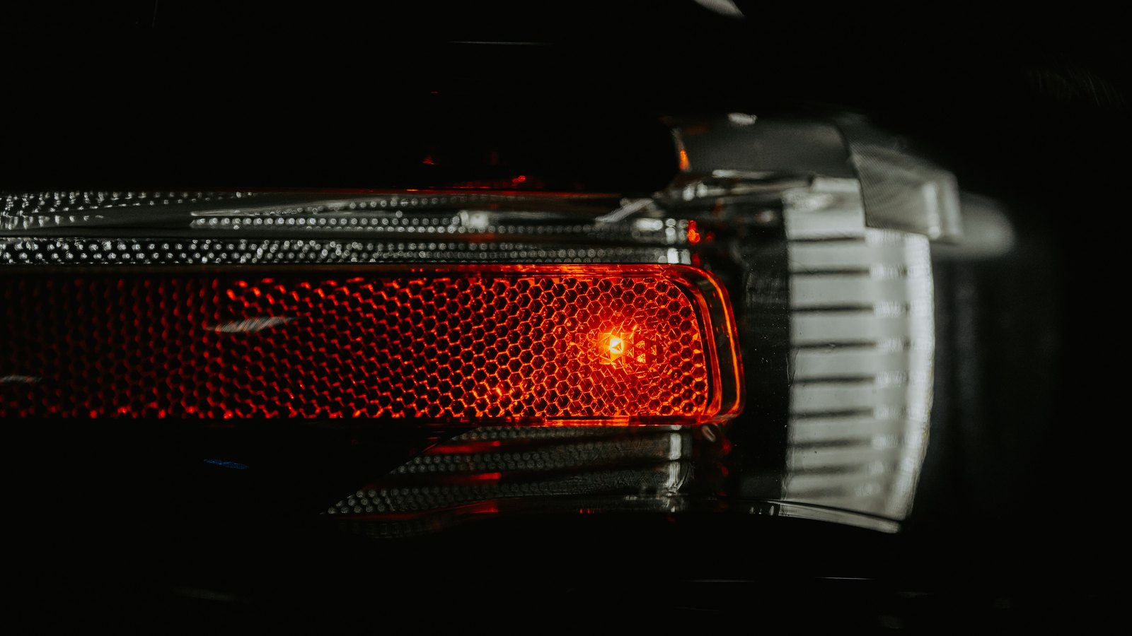 A tight night-time close up of the red brake light of a car, its honeycomb textured lens glowing against near total darkness, with a chrome trim strip and the edge of a clear indicator lens beside it. No registration plate, person, road sign, lettering, agency marking, vehicle badge or identifiable place is visible anywhere in the frame. A generic view of a vehicle braking in the dark, not a photograph of this event.