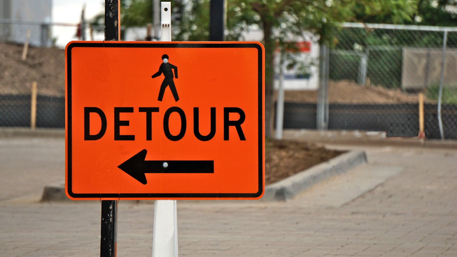 A standard orange detour sign with a black pedestrian icon and a left-pointing arrow, mounted kerbside beside a temporary fence. No place name, no brand, no person, no vehicle and no number plate appears in the frame. A stock photograph, not a photograph of this event.
