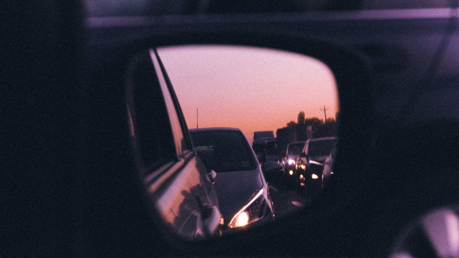 A view through a car's wing mirror at dusk showing a queue of stopped vehicles on the road behind, headlights on, against an orange and violet sky above a low, indistinct treeline. No registration plate, road sign, lettering, landmark or identifiable person is legible anywhere in the frame. A generic illustration of traffic held up behind a closure, not a photograph of this event.
