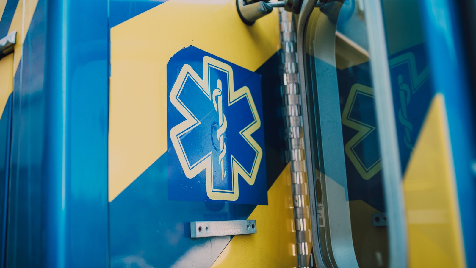A close view of the side door of an ambulance, painted in blue and yellow blocks, carrying the six pointed star of life emblem with its staff and serpent in the centre, beside a window frame and a hinge strip. No lettering, agency name, registration plate, person or place is visible anywhere in the frame. A generic illustration of an emergency medical vehicle, not a photograph of this event.