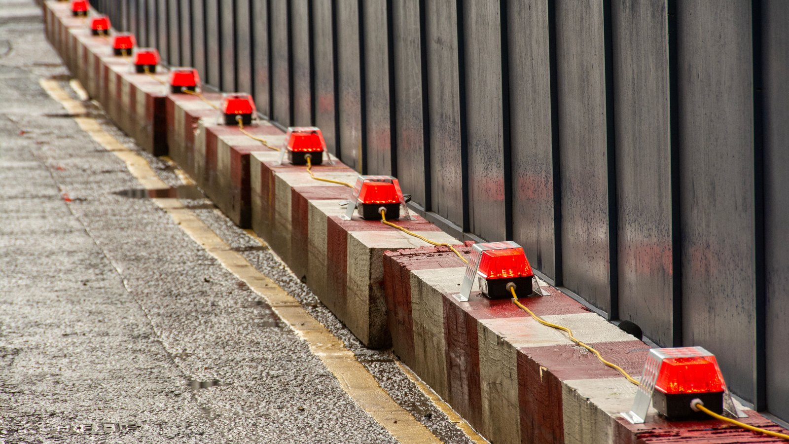 A row of red warning lamps bolted along the top of a red and white striped concrete barrier, their wiring looped between them in a yellow cable, running away from the camera beside a wet concrete roadway with a painted white edge line. A ribbed steel wall stands behind the barrier. No person, vehicle, registration plate, road sign, agency marking or identifiable place is visible in the frame. A generic view of warning lights marking off a closed section of roadway, not a photograph of this event.