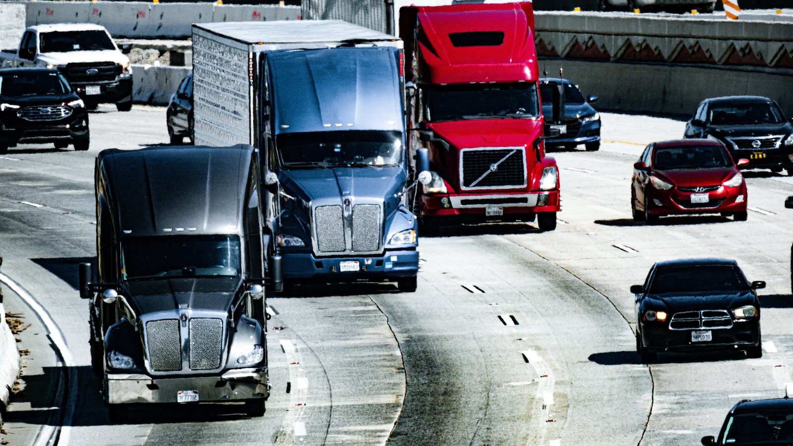 Two dark tractor units and a red one queue nose to tail in the left lanes of a multilane divided highway in bright sunlight, with saloon cars and a pickup moving in the lanes to the right, a concrete barrier wall and a chevron marked crash cushion along the far shoulder, and no legible number plates, signs, agency markings or place names anywhere in the frame. A generic illustration of traffic on an interstate carriageway, not a photograph of this event.