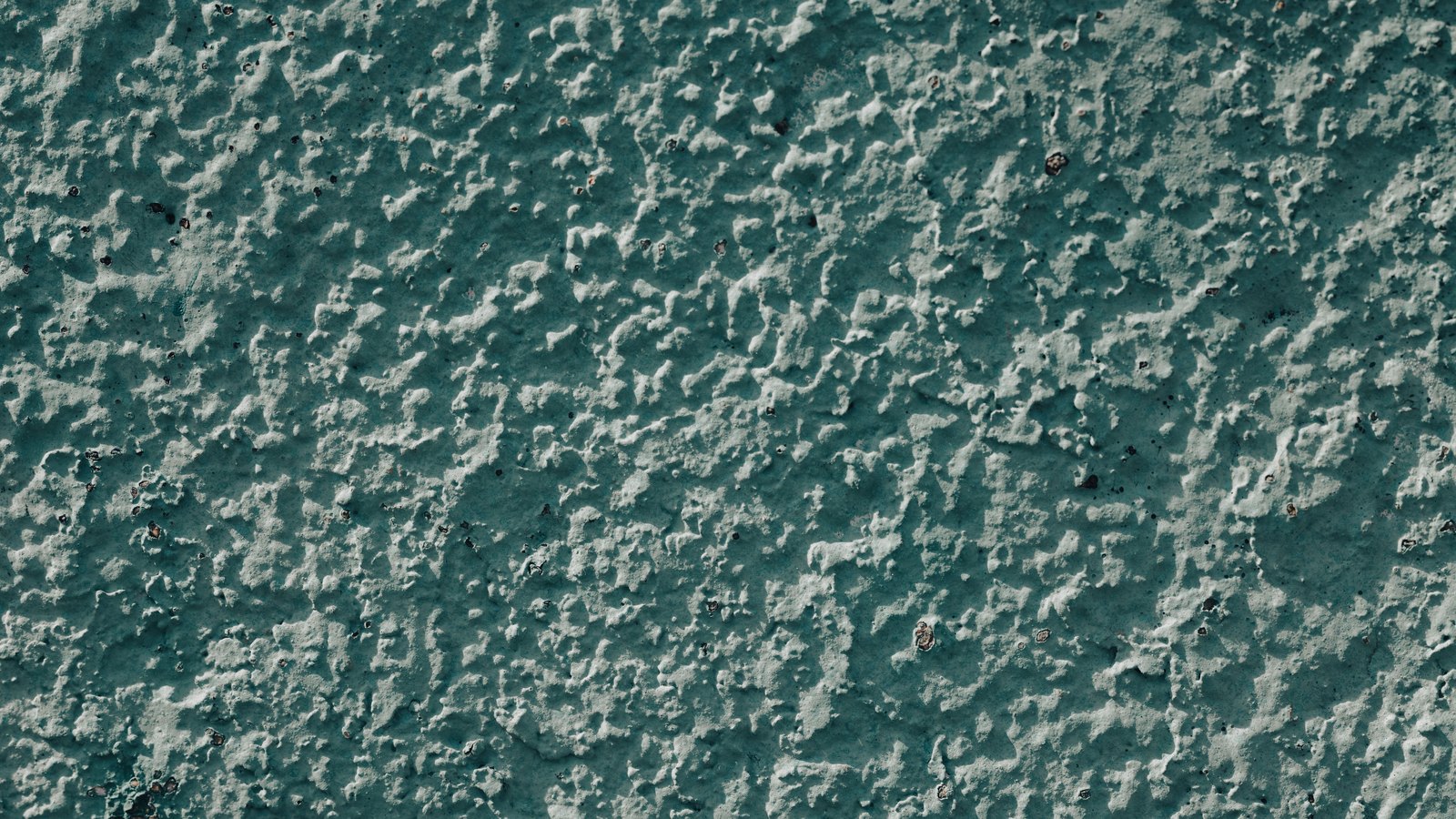 A close crop of a rough textured rendered wall painted a muted blue green, the raised grain of the render catching the light across the whole frame. No text, signage, route marker, lettering, vehicle, person, emergency service or identifiable place is visible. A plain surface illustration, not a photograph of Interstate 75, of mile marker 140 or of the vehicle fire, and not evidence about the condition of any road.