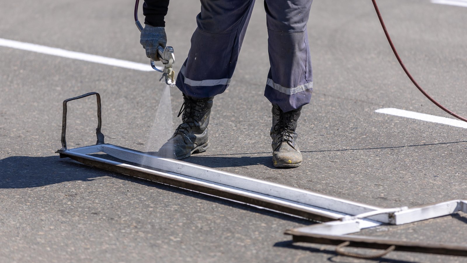 Fresh white lane markings on grey asphalt, with a long metal stencil rail laid along the surface and a road worker seen only from the waist down in plain navy trousers and scuffed boots, holding a paint sprayer. No face, no vehicle, no number plate, no signage, no agency marking and no legible text of any kind appears in the frame. A generic illustration of highway lane markings, not a photograph of this event or this road.