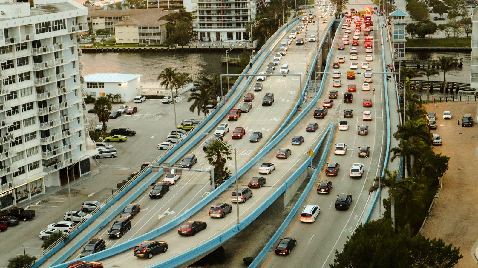 Dense traffic fills every lane of a multi-lane road as it rises onto a bridge in a Florida coastal city, with cars, pickups and vans queued nose to tail on both carriageways, palm trees and low white apartment blocks along the water on either side, and no legible number plates, road signs, agency markings or place names anywhere in the frame. A generic illustration of congested traffic on a Florida road, not a photograph of this event.