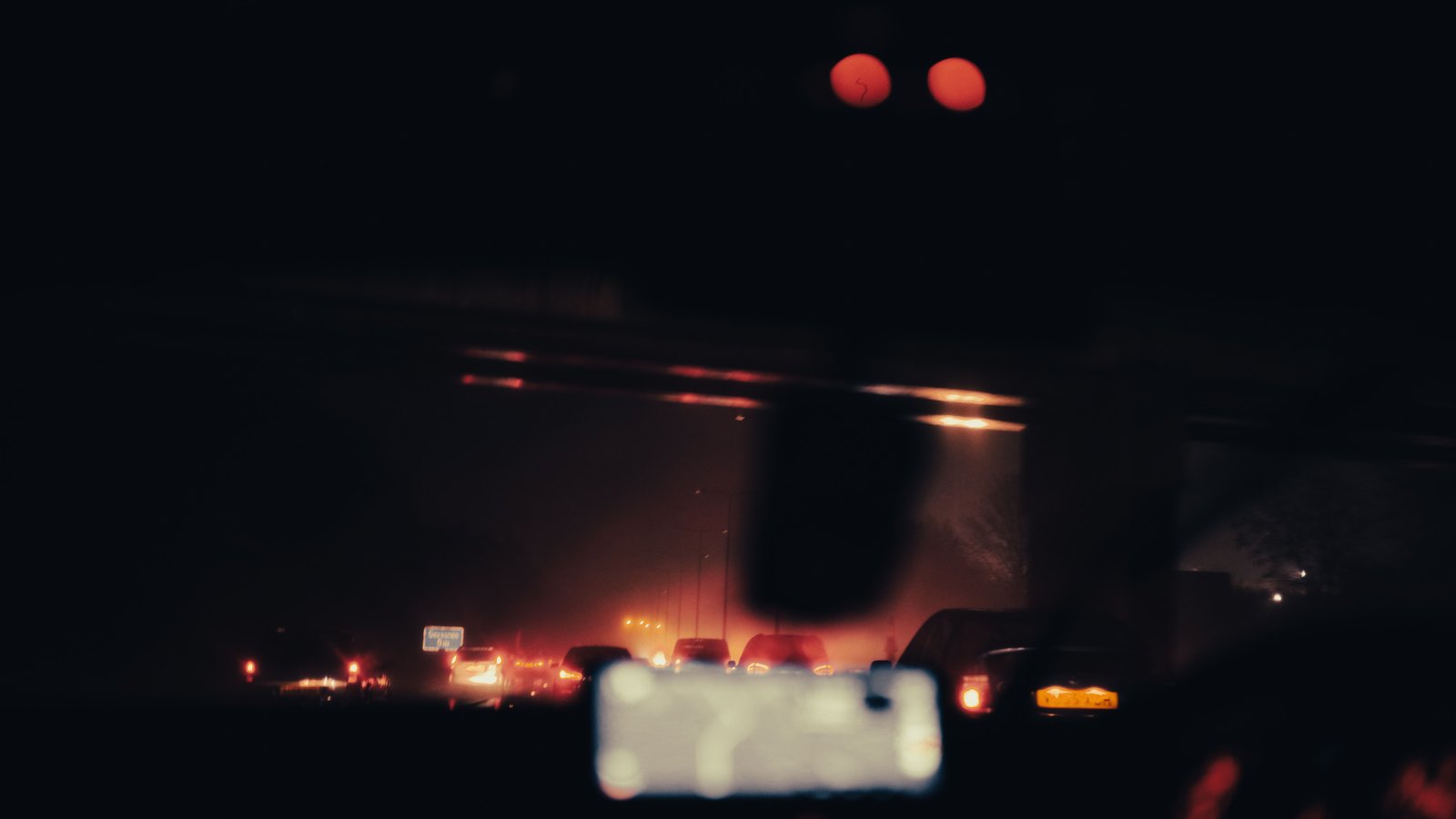 A dark, deliberately blurred view forward through a car windscreen at night on a multi lane road, with the red tail lights of slowed traffic strung out ahead and an overhead sign gantry silhouetted against the sky. No lettering, number plate, vehicle marking or landmark is legible anywhere in the frame. It is a stock photograph of night traffic and not a photograph of this event.