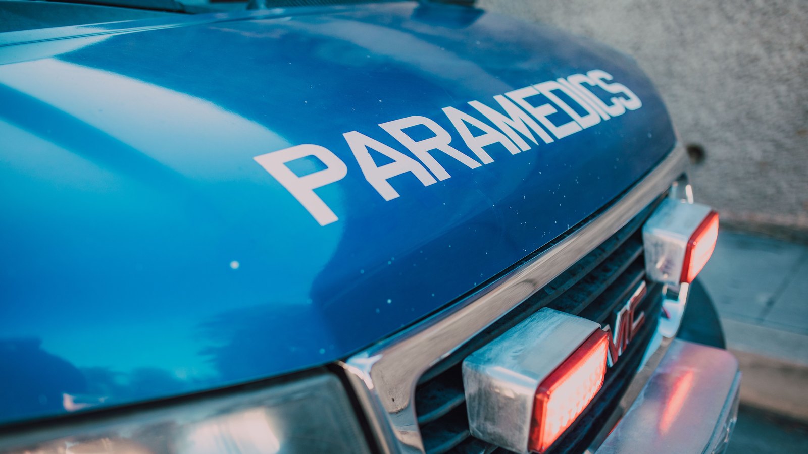 The blue bonnet and chrome grille of an American ambulance fill the frame, with the single word PARAMEDICS lettered in white across the bonnet and two red warning lamps lit in the grille. The only other lettering is the vehicle maker's badge. No people, no number plate, no street, no building, no place name and no department or agency marking appear anywhere in the frame. Stock photograph, not a photograph of this event.