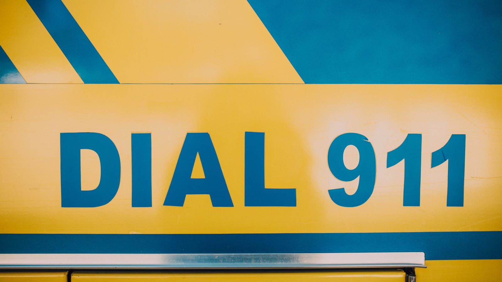 A tight close-up of the flank of an emergency vehicle, painted yellow with blue diagonal stripes and carrying the words DIAL 911 in large blue block letters, with a metal grab rail running along the bottom of the frame. No agency name, department, unit number, licence plate, person or place is visible anywhere in the picture. A generic illustration of emergency response, not a photograph of this event.