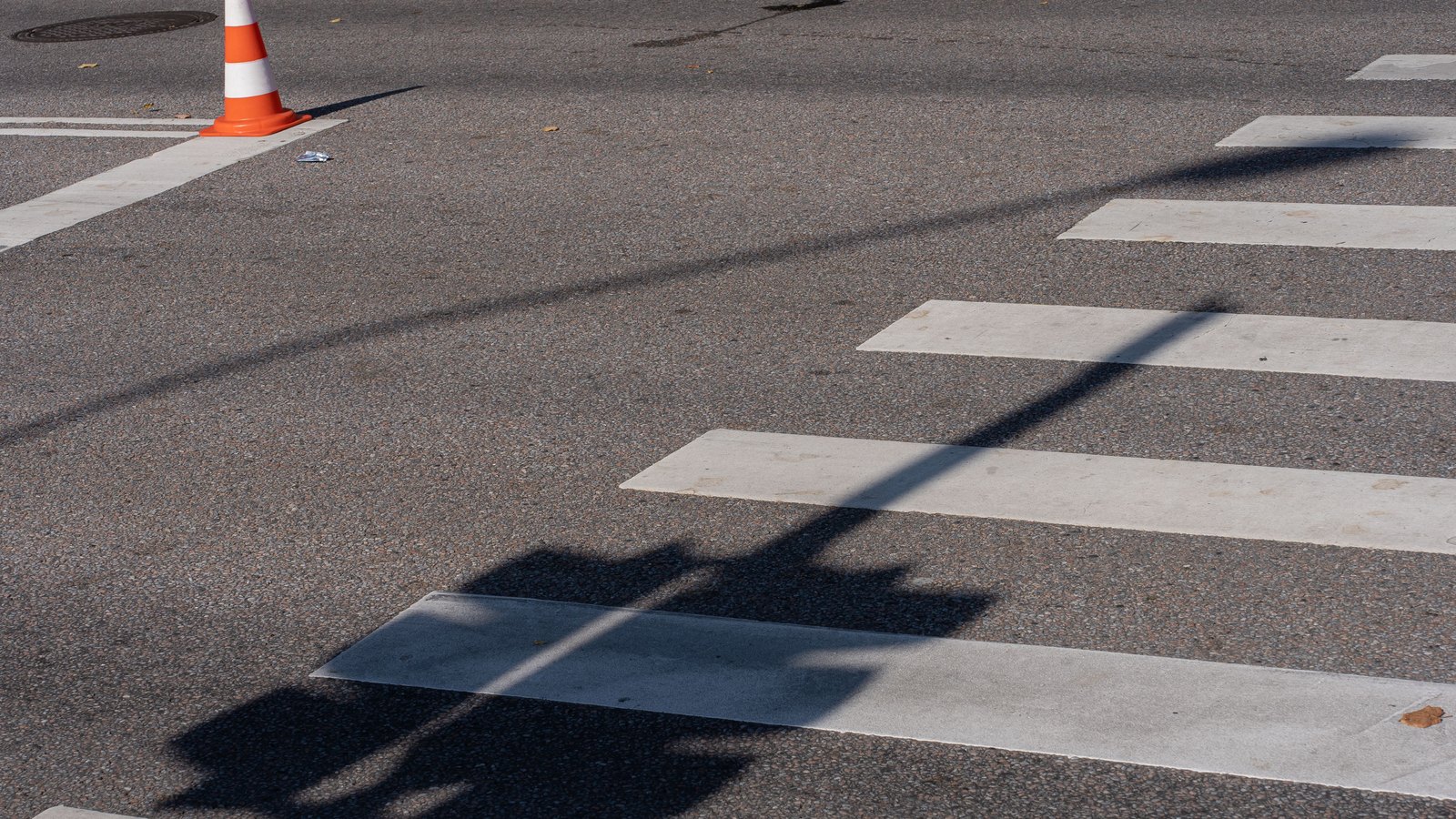 A single orange and white traffic cone stands on sunlit grey asphalt at the edge of a painted white crosswalk, with the long shadow of a traffic signal falling across the road surface. No people, vehicles, number plates, signs, agency markings or place names appear anywhere in the frame. Stock photograph, not a photograph of this event.