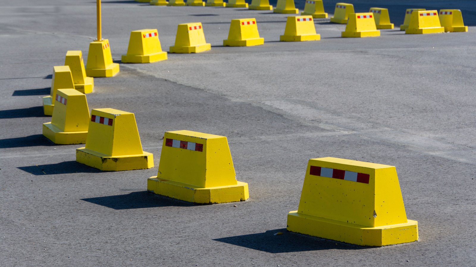 A line of low yellow concrete traffic barricades, each with a red and white reflective band on its side, set out across bright sunlit grey asphalt in two curving rows, with a thin yellow post and a blue metal railing behind them, and no people, vehicles, number plates, signs, text or brand marks anywhere in the frame. A generic illustration of traffic control equipment, not a photograph of this event.