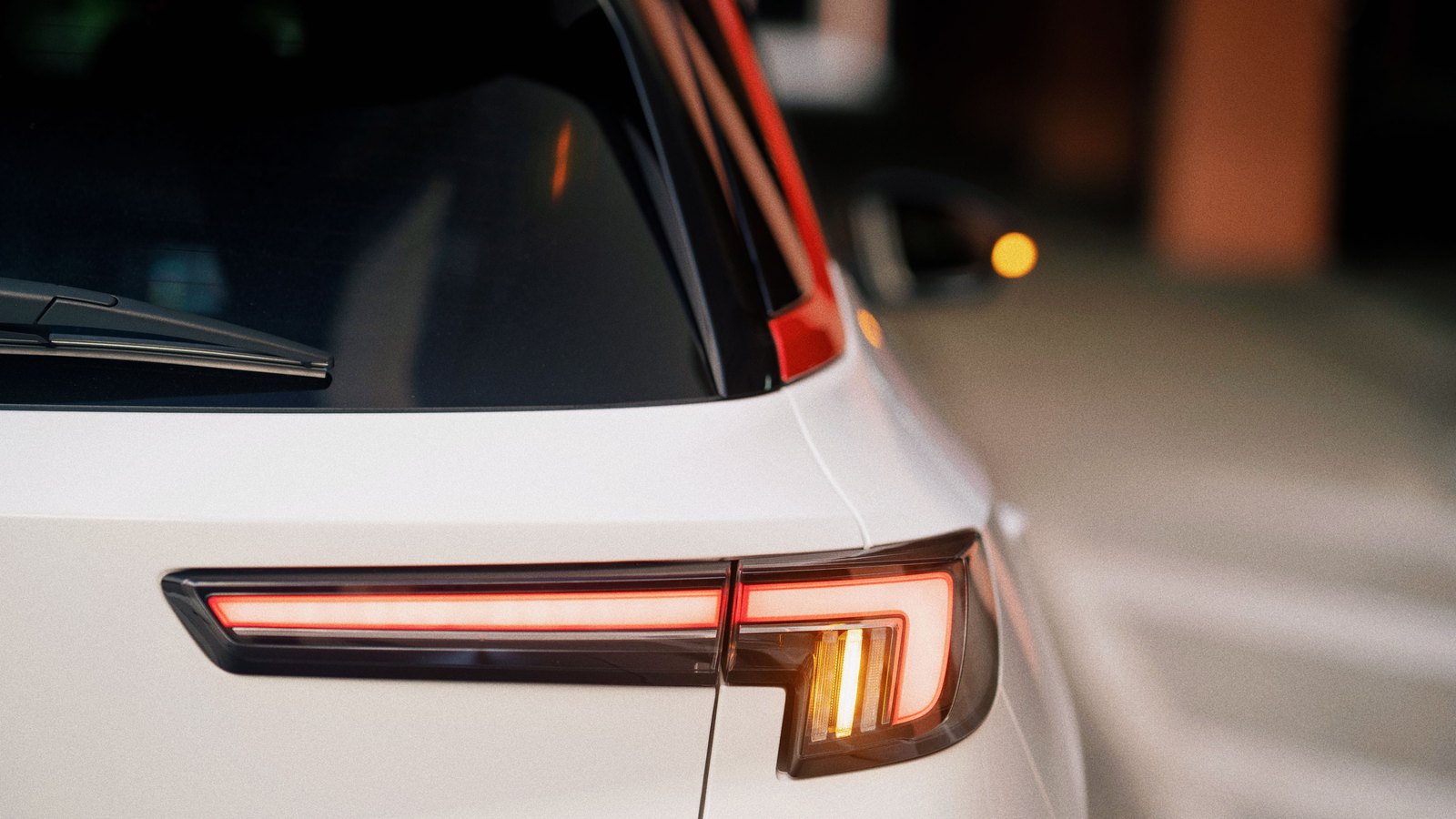 A tight close-up of the rear light cluster of a white car, the brake strip glowing red across the tailgate and the amber indicator lit beside it, the background thrown out of focus behind. No person, no readable licence plate, no agency name or livery, no signage and no identifiable place appears in the frame. A generic illustration of a stopped vehicle showing its lights, not a photograph of this event.