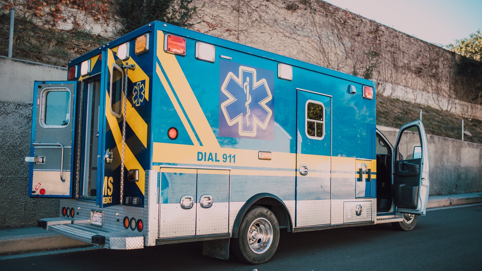 A blue and yellow ambulance photographed from behind and to one side, parked on a paved roadside in daylight with its rear and side doors standing open, a star of life emblem on the body and the words DIAL 911 along the flank. A bare earth bank rises behind it. No agency name, department, unit number, readable licence plate, person or identifiable place appears anywhere in the frame. A generic illustration of roadside emergency response, not a photograph of this event.