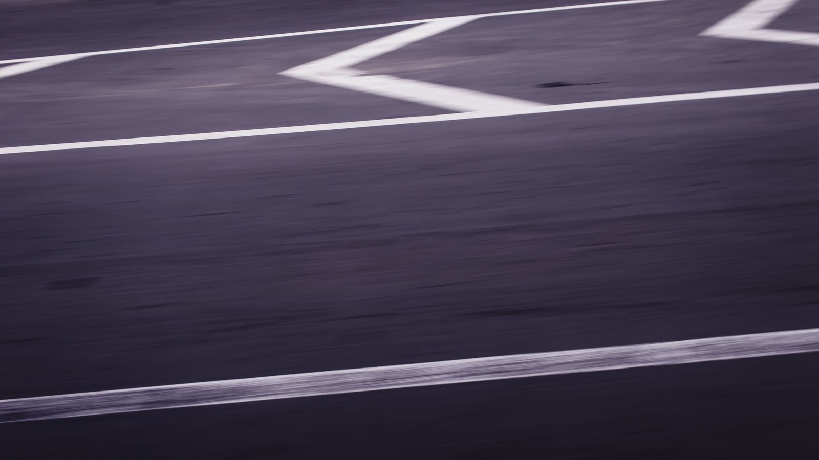 A motion blurred close crop of dark grey asphalt carrying three white painted lines and part of a white chevron, shot from a moving vehicle so the surface streaks horizontally. No vehicles, people, signage or lettering are visible and no place can be identified from it. An abstract road surface used as a plain illustration, not a photograph of Interstate 75, of North Fort Myers or of this crash, and not evidence about the condition of any road.