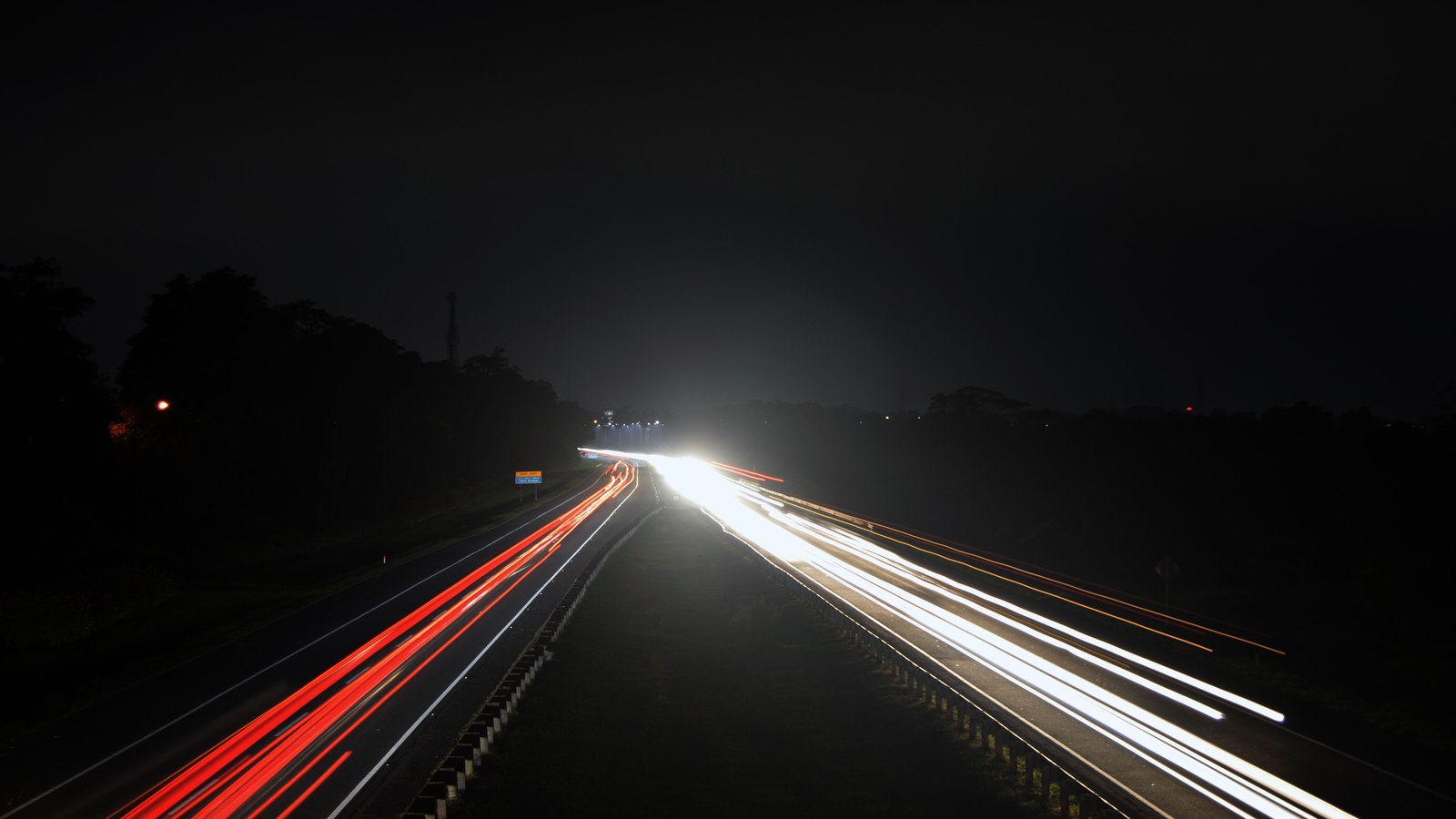 A long exposure looking down a flat divided highway at night. Red tail light trails run away from the camera on the left carriageway and white headlight trails run toward it on the right, separated by a low guard rail, with a dark treeline on both verges and no legible sign, marking, number plate or landmark anywhere in the frame. Not a photograph of this event.