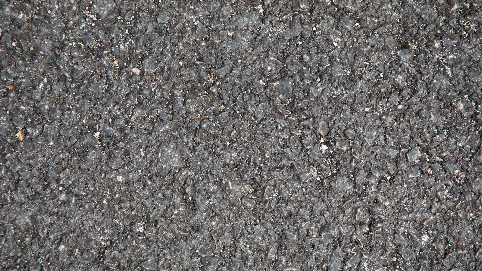 A close overhead view of coarse asphalt filling the frame, its dark aggregate flecked with pale grey and white stone.