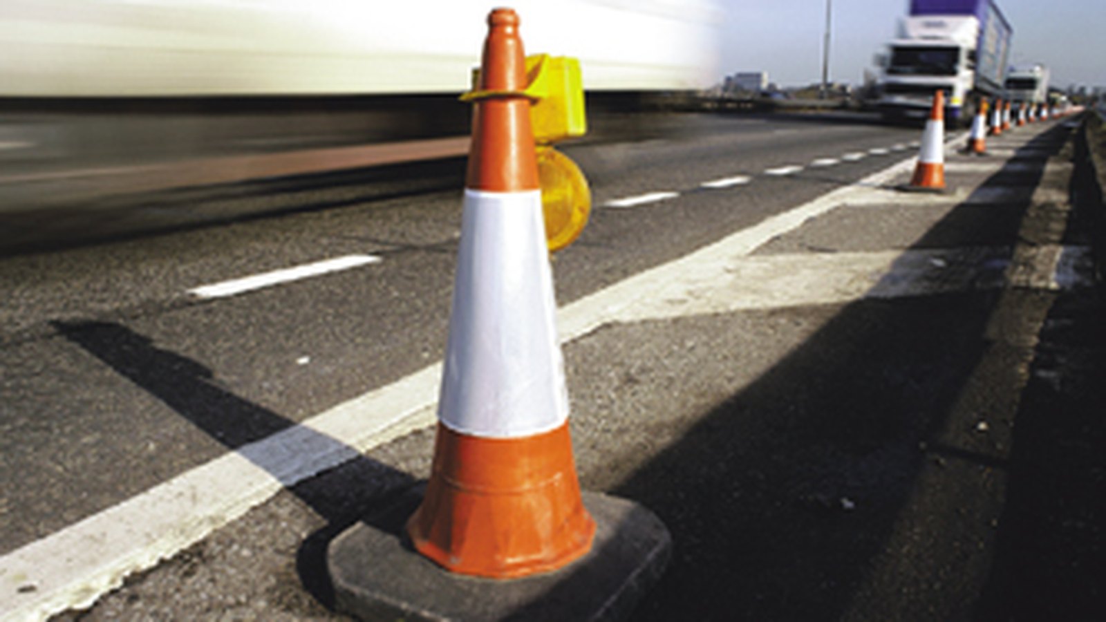 An orange traffic cone with a reflector beside a highway work zone and blurred trucks, not a photograph of this event.