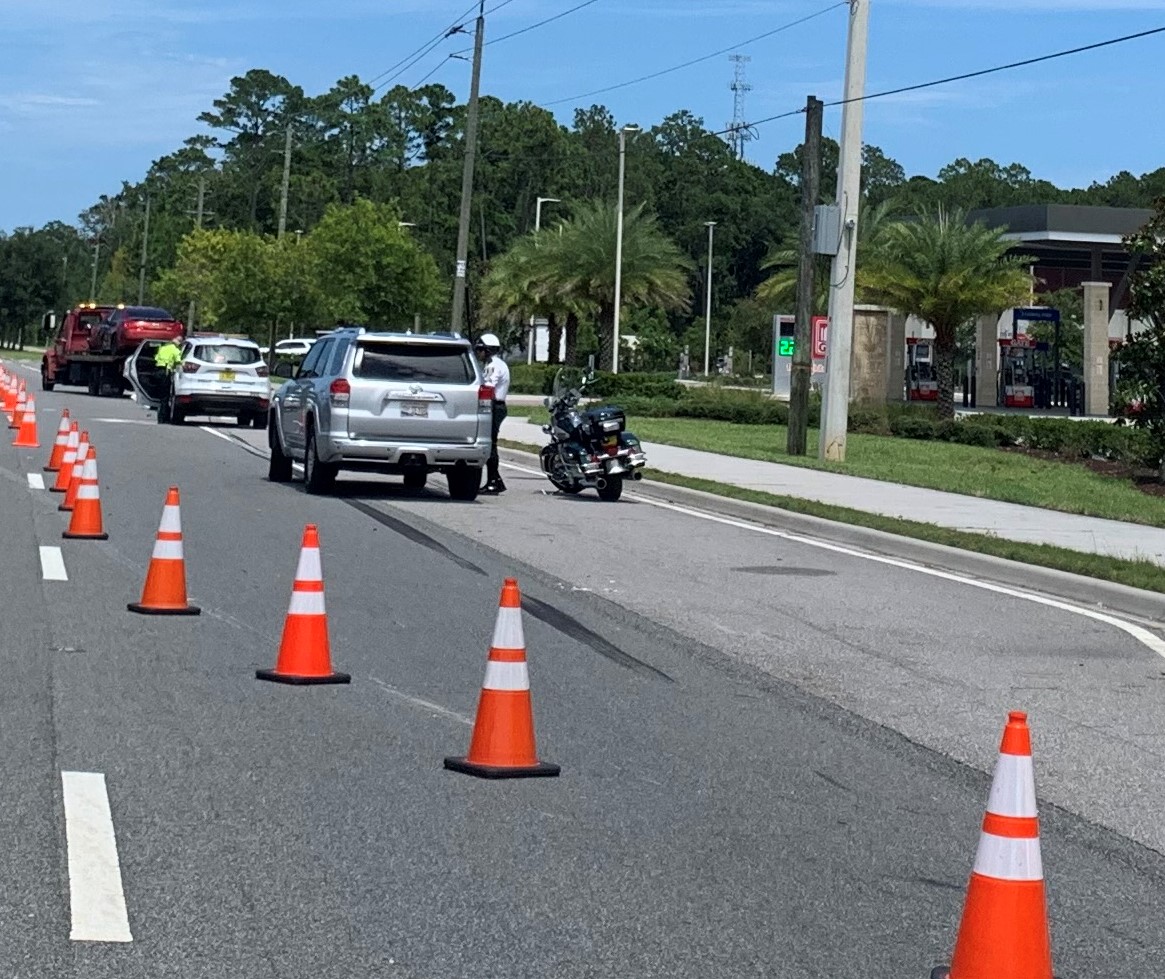 Orange traffic cones channel vehicles past an incident scene on a palm-lined road, not a photograph of this event.