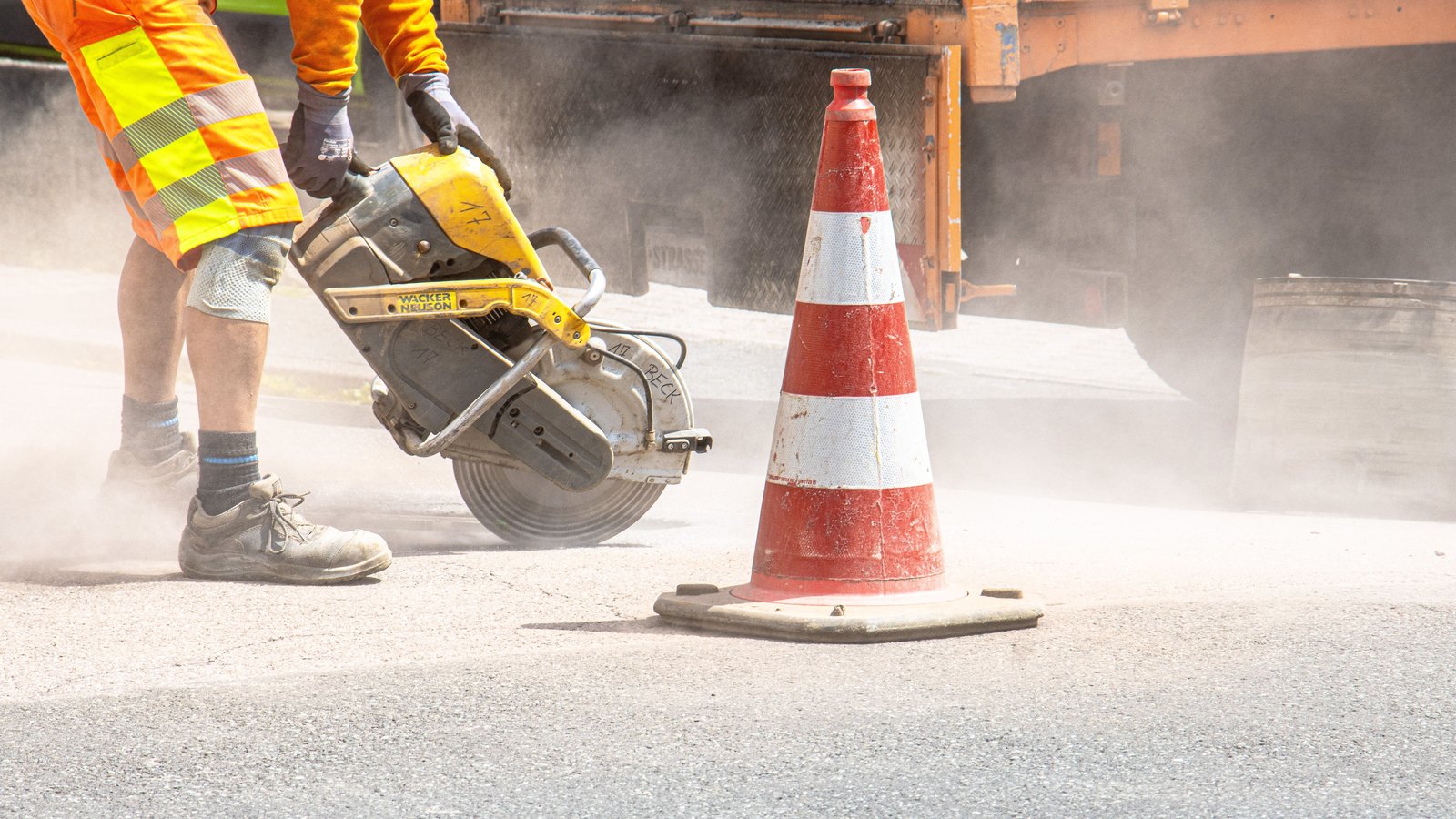 An orange traffic cone beside a road worker and pavement-cutting equipment.