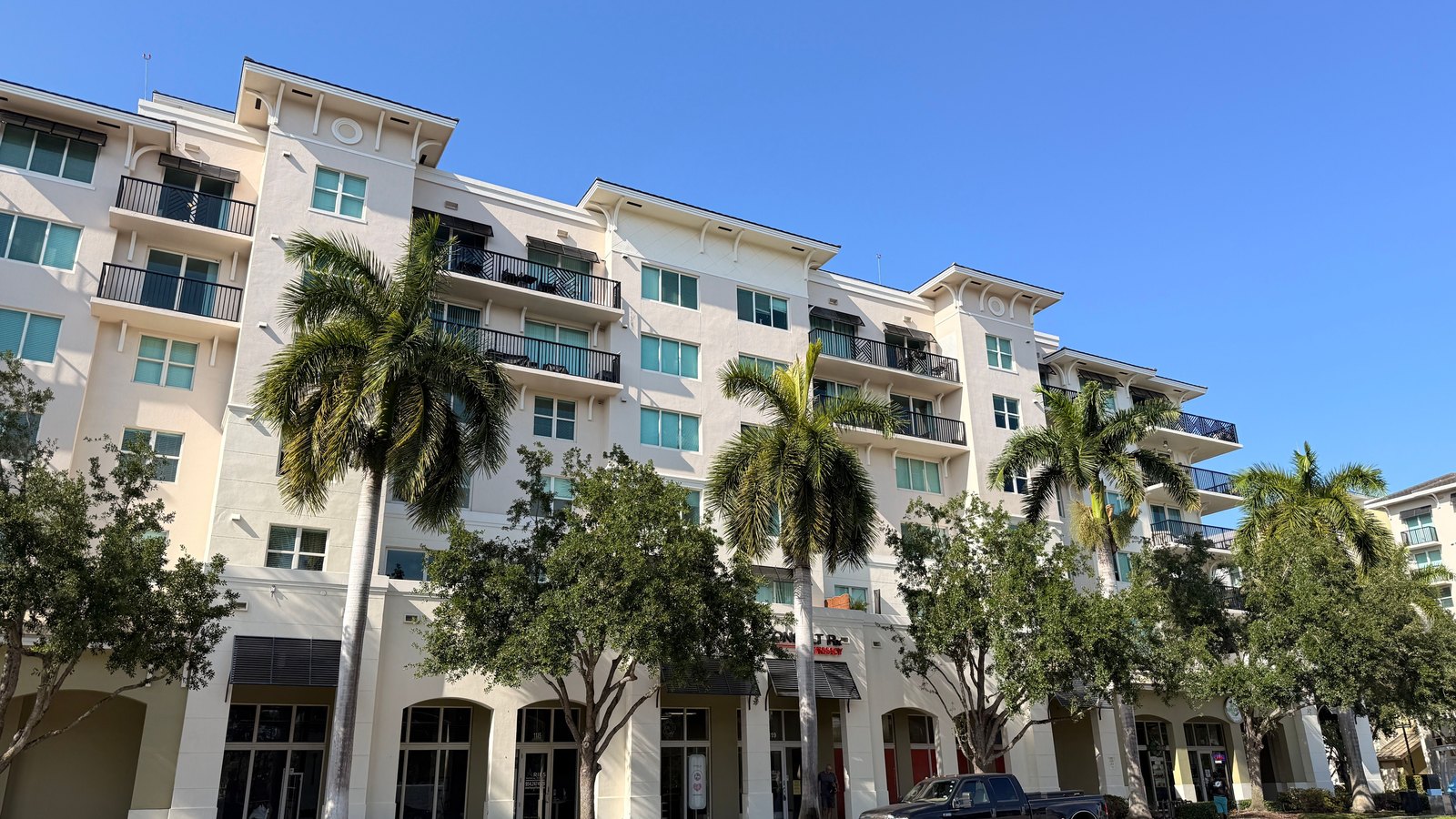 A five storey cream stucco apartment building with balconies, seen from across a road in flat midday sunshine under a clear blue sky, with young palms and shade trees along the pavement and a single dark pickup truck parked at the kerb. No people are in the frame and no street name, address number or number plate is legible. A generic illustration of a Florida residential community, not a photograph of this event.
