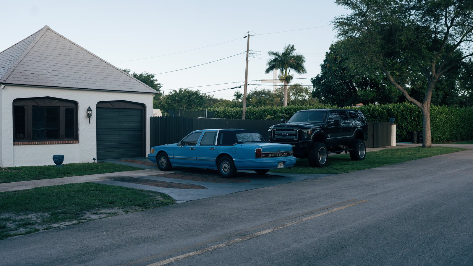 A quiet residential street in soft evening light, with a single storey white stucco house and a closed garage door on the left, two vehicles parked on the driveway beside it, a trimmed hedge and palms behind, and flat ground running back to the treeline. No people appear, and there is no street number, no signage, no agency marking and no legible number plate anywhere in the frame. A generic illustration of a Southwest Florida residential street, not a photograph of this event.
