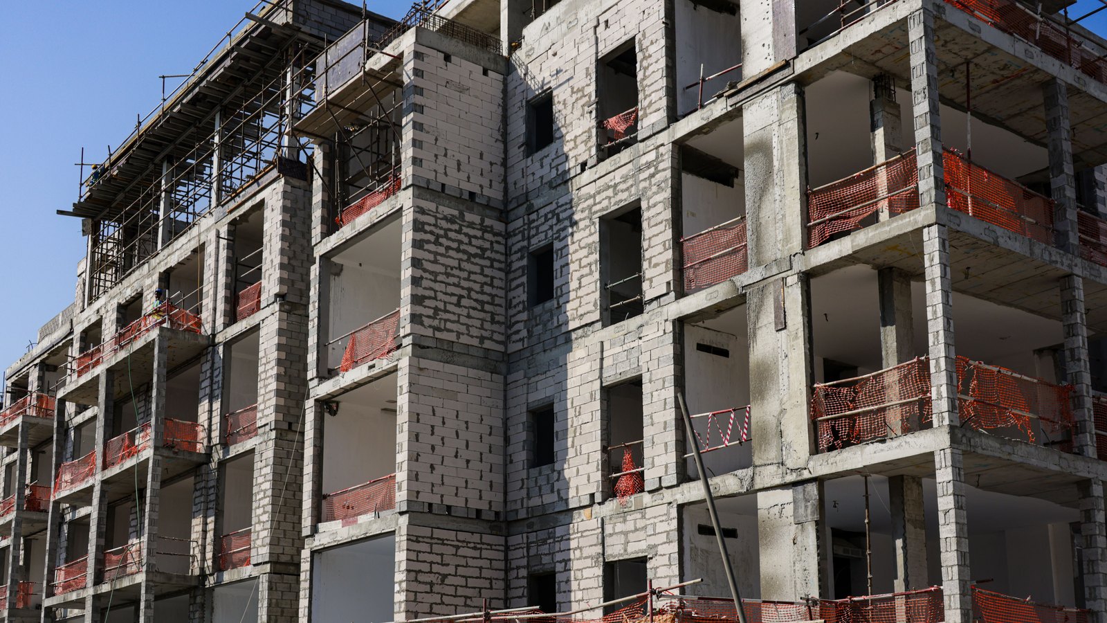 An unfinished multi-story concrete apartment building with open window bays, block walls, scaffolding and orange safety netting under a clear blue sky. No person, sign, logo, address or other identifying detail is visible. Stock photo, not a photograph of this event.