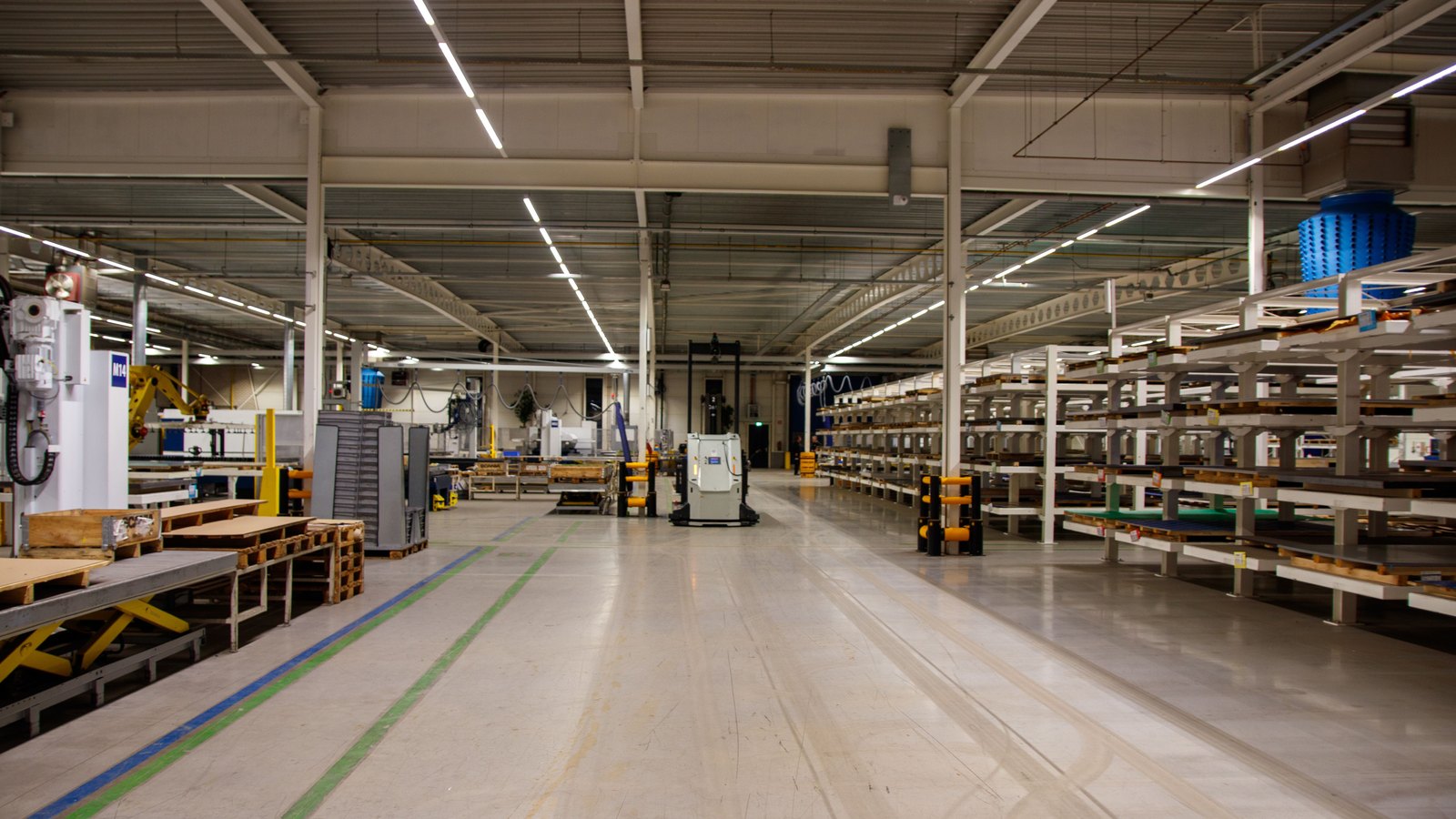 The inside of a quiet manufacturing hall lit by rows of fluorescent strips, with a wide empty aisle running down the centre marked by blue and green floor tape, steel racking stacked with sheet material along the right, work benches and a yellow robot arm along the left, and a small white electric tug parked in the middle distance. Nobody is close enough to identify and no company name is legible.
