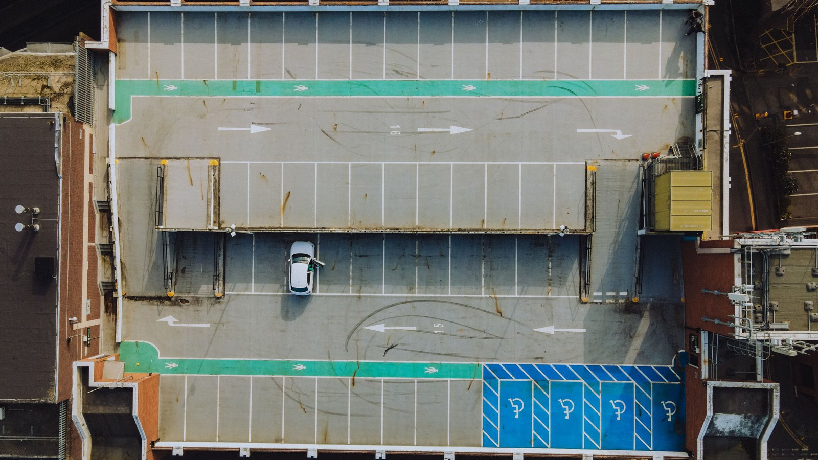 A rooftop car park photographed from directly overhead, almost entirely empty, with white painted bays, direction arrows and a green pedestrian lane across it. One white car stands in a bay near the middle. No signage, brand mark or person is in the frame.