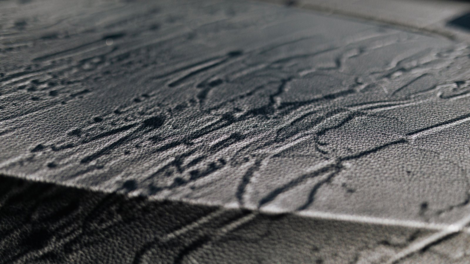 A close photograph of black leather lit from a low angle, the pebbled grain running across the whole frame and broken by long shallow creases that catch the light as pale lines. No text, lettering, brand mark, object, person or place is in view.