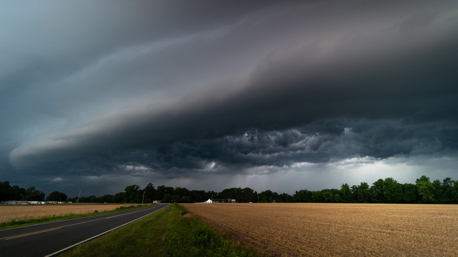 Dark shelf cloud over a flat rural road and fields, not a photograph of this event.