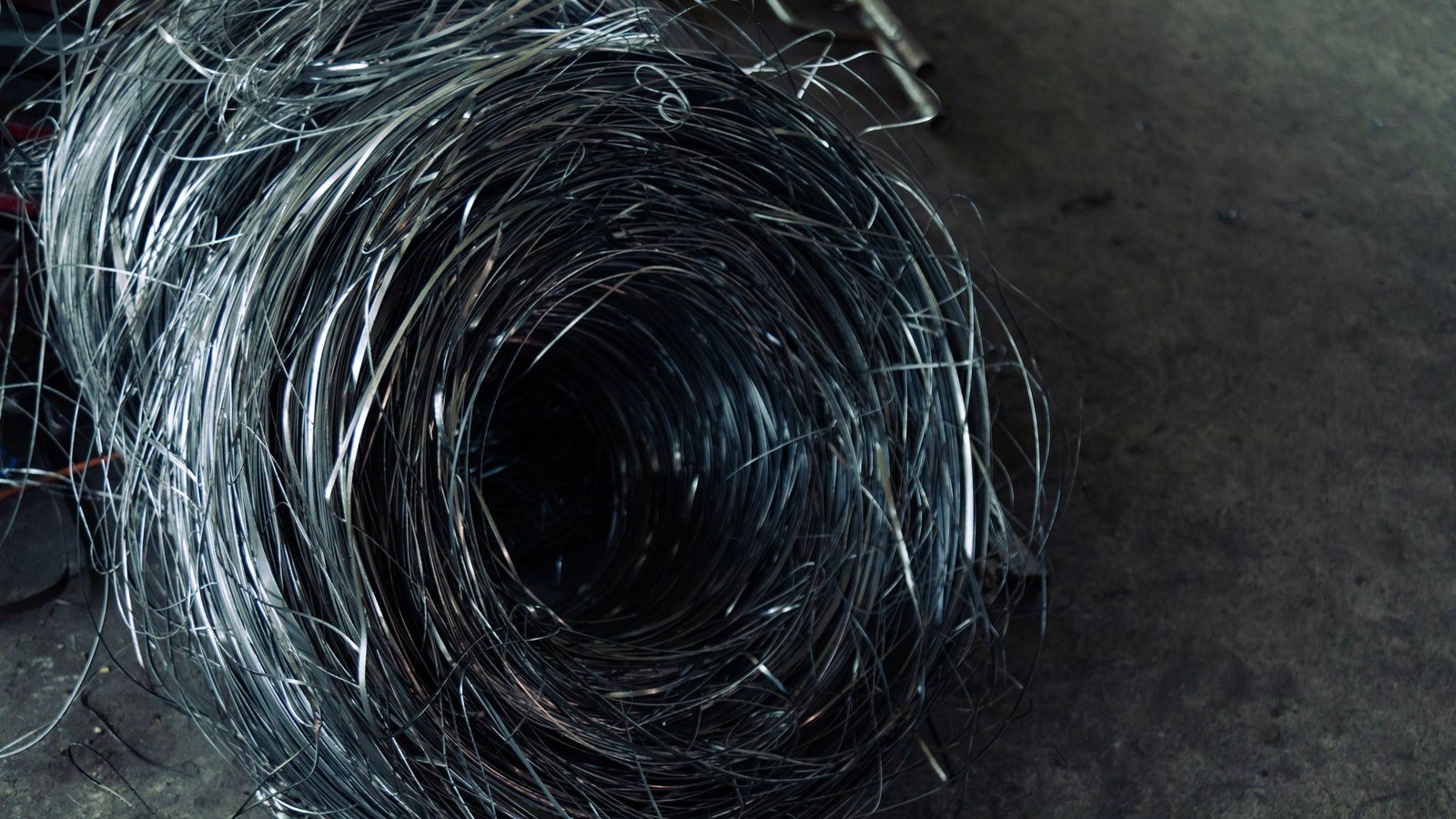 A large loose coil of bright steel wire lying on a plain concrete floor, the strands spiralling inward to a dark centre, with stray ends standing out from the mass. No person, brand, lettering or place is in the frame.