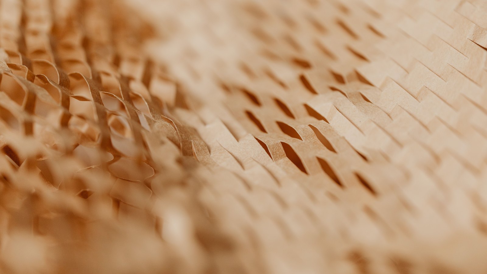 A close photograph of honeycomb kraft packaging paper, warm tan slit-cut sheets pulled open into staggered rows of hexagonal cells that recede out of focus toward the left of the frame. No text, lettering, brand mark, product, tool, person or place is in view.