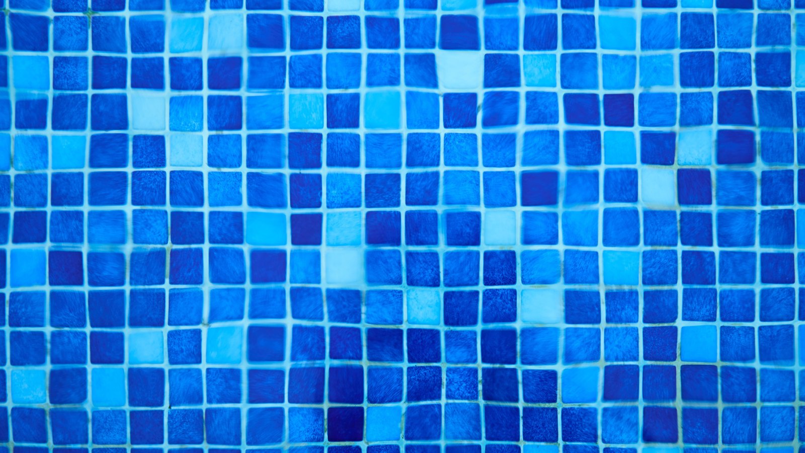 A tight overhead crop of small square tiles in many shades of blue, from pale turquoise to deep navy, laid in an even grid with fine pale joints between them and seen through shallow water that bends the lines slightly. The pattern fills the whole frame. No text, brand mark, person or place is in view.