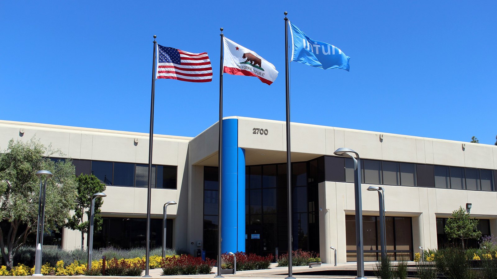 The entrance to 2700 Coast Avenue in Mountain View, California, a low cream and grey office building with the number 2700 above the door and a blue clad column beside it, three flagpoles on the forecourt carrying the American flag, the California state flag and a pale blue flag carrying the company name in white, under a clear sky.