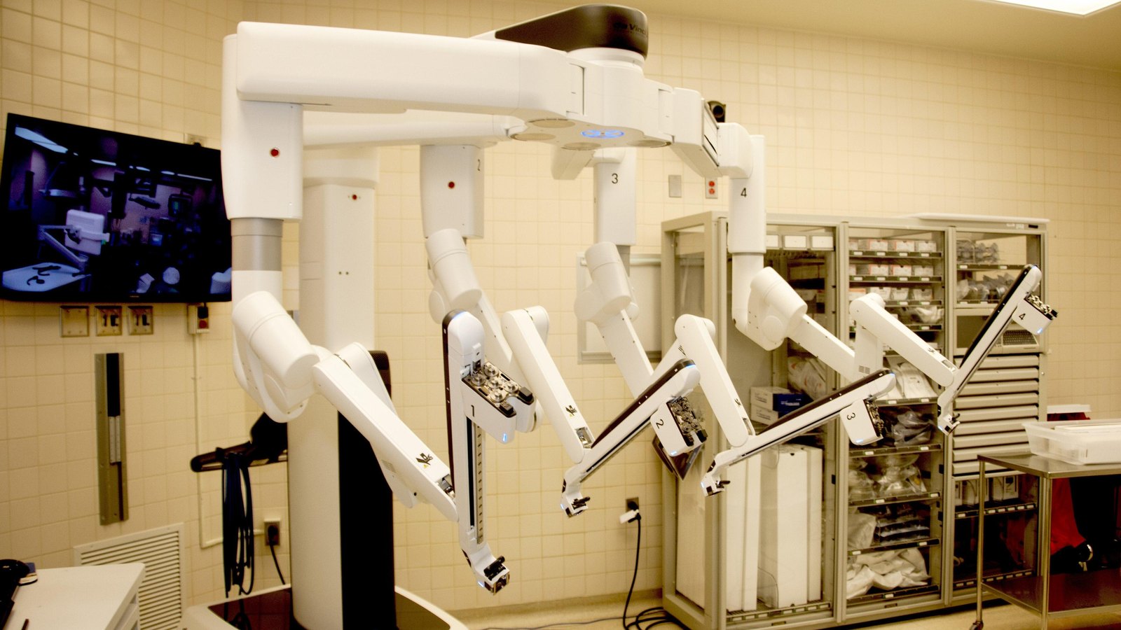 A white da Vinci Xi surgical robot with four numbered arms extended in an empty operating room at William Beaumont Army Medical Center, with a wall monitor at left and supply cabinets behind.