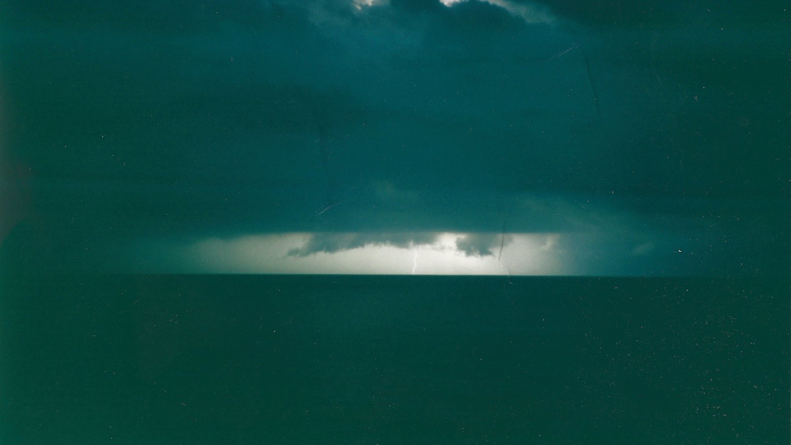 A dark expanse of open ocean at night beneath a heavy cloud deck, with a bright band of distant thunderstorm light and a faint lightning strike glowing along the horizon. No land, vessel, person, structure, signage or marking of any kind appears anywhere in the frame. A generic illustration of the kind of thunderstorm activity a tropical disturbance produces over open water, not a photograph of this event.