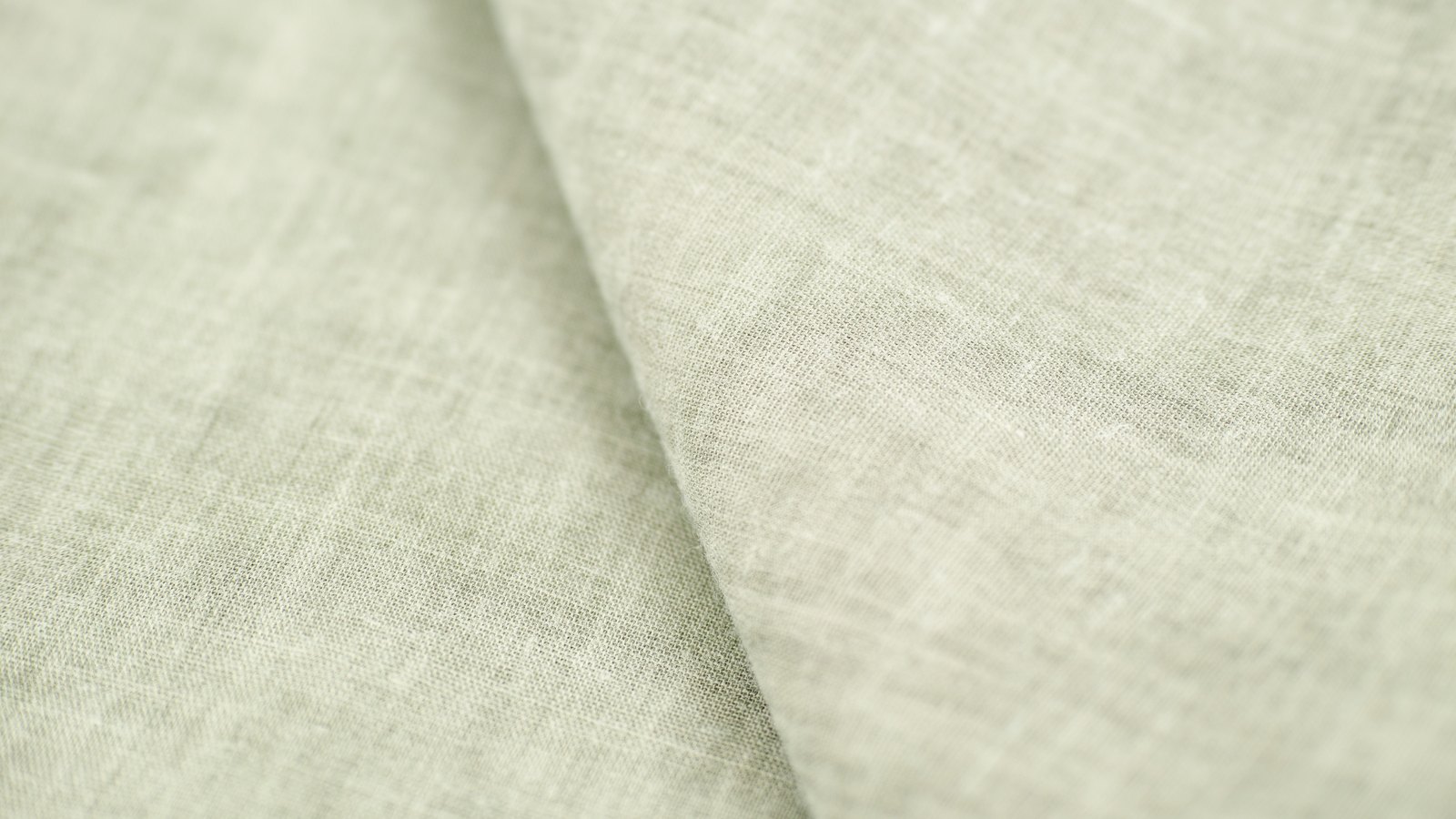A close photograph of pale sage-grey woven linen cloth filling the frame and falling into a single soft vertical fold, the weave visible across the surface, with no pattern, label or lettering.