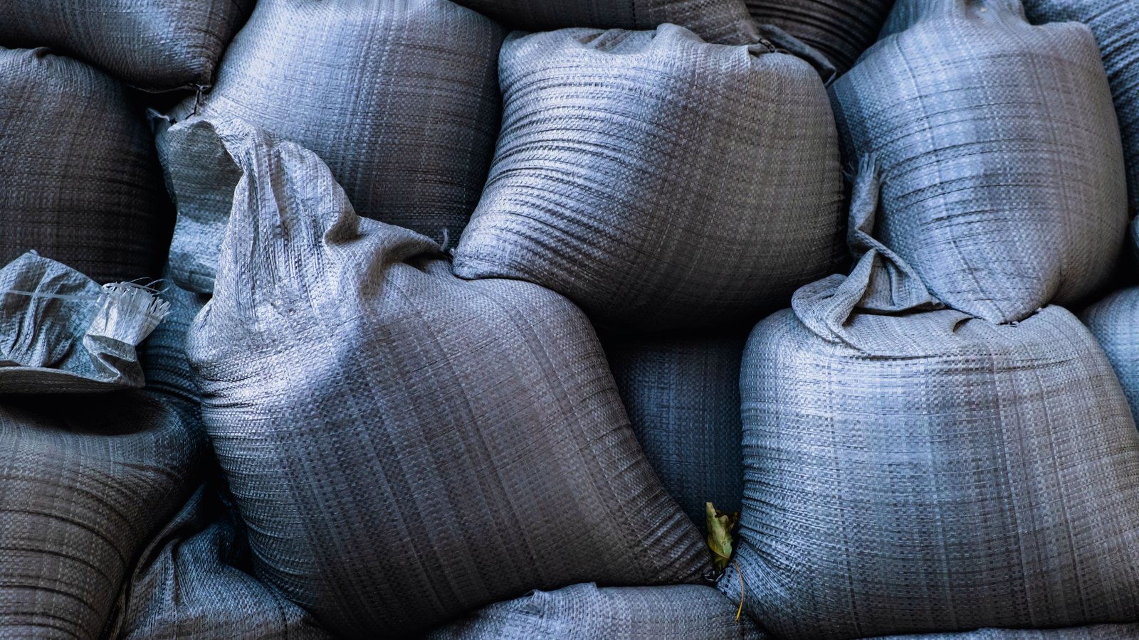 A tightly stacked wall of grey woven sandbags filling the frame, one bag torn open at the left edge.