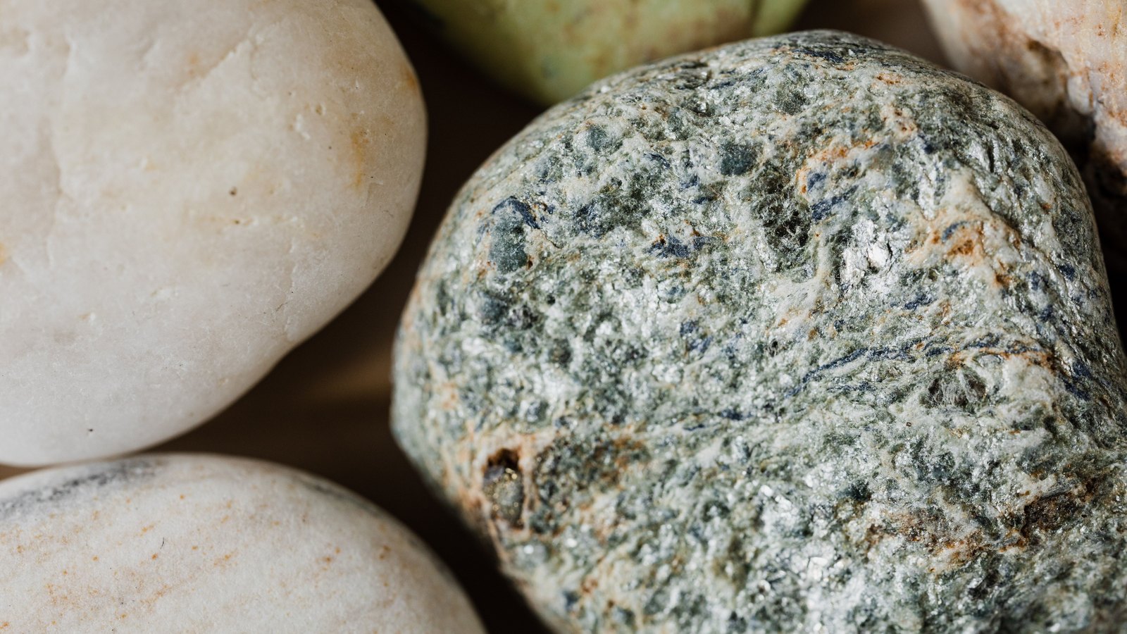 A close photograph of smooth rounded pebbles packed together and filling the frame. The largest, right of centre, is a mottled grey green stone flecked with darker green, blue grey and rust and scattered with bright mica glints, and around it sit three cream white pebbles and one pale green one. No text, marking, brand mark, person or horizon is in view.