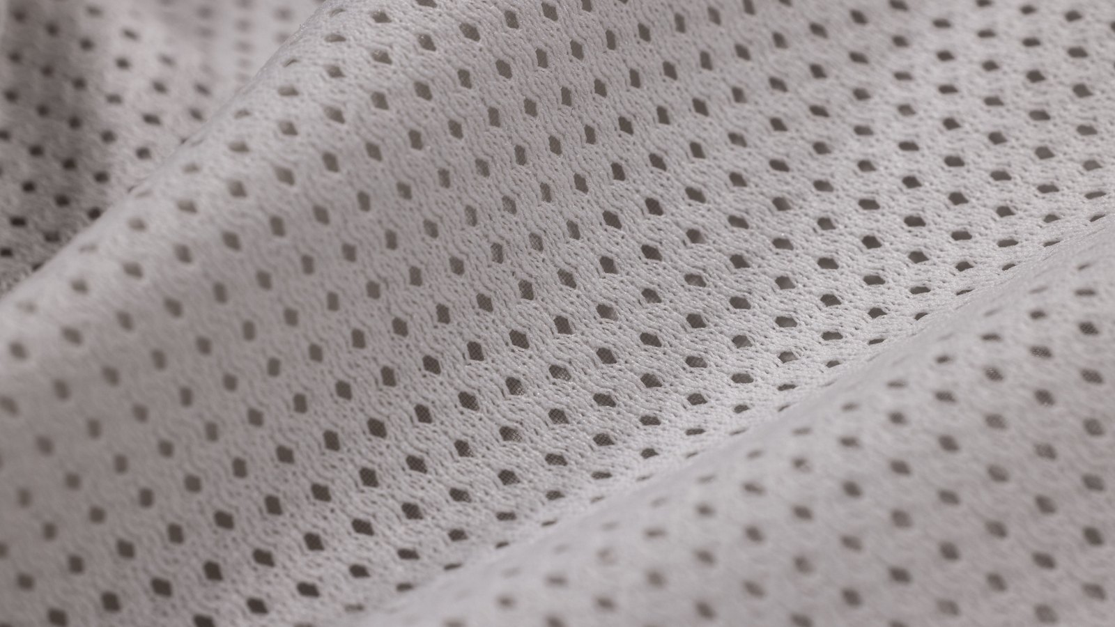 A close photograph of white open-mesh knitted fabric filling the frame, its regular grid of small perforations running diagonally across two soft folds, the darker shadow beneath showing through every hole.