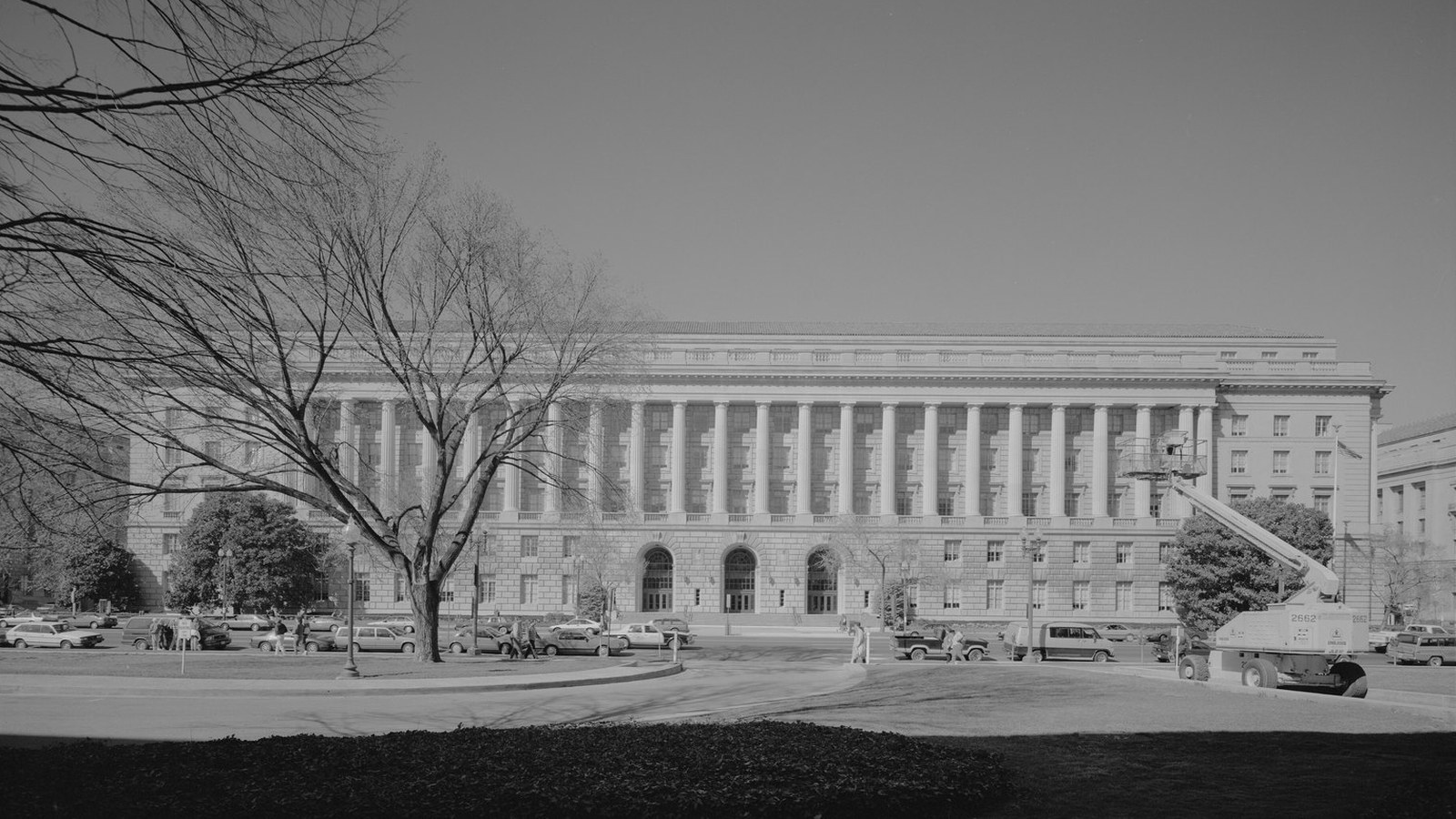 The Constitution Avenue front of the Internal Revenue Service headquarters in Washington, a long colonnade running the width of the building above an arched entrance, photographed in black and white with bare trees and parked cars in front.