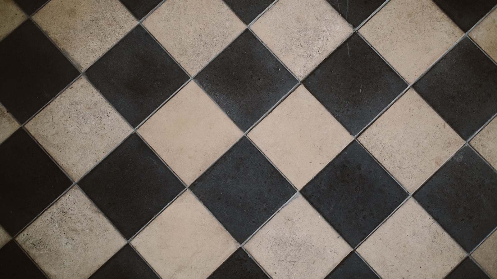 A square-on overhead crop of a floor of large chequerboard tiles set on the diagonal, alternating matt near-black and pale cream, the surfaces scuffed and faintly stained with age. The pattern fills the frame. No text, brand mark, person or place is in view.