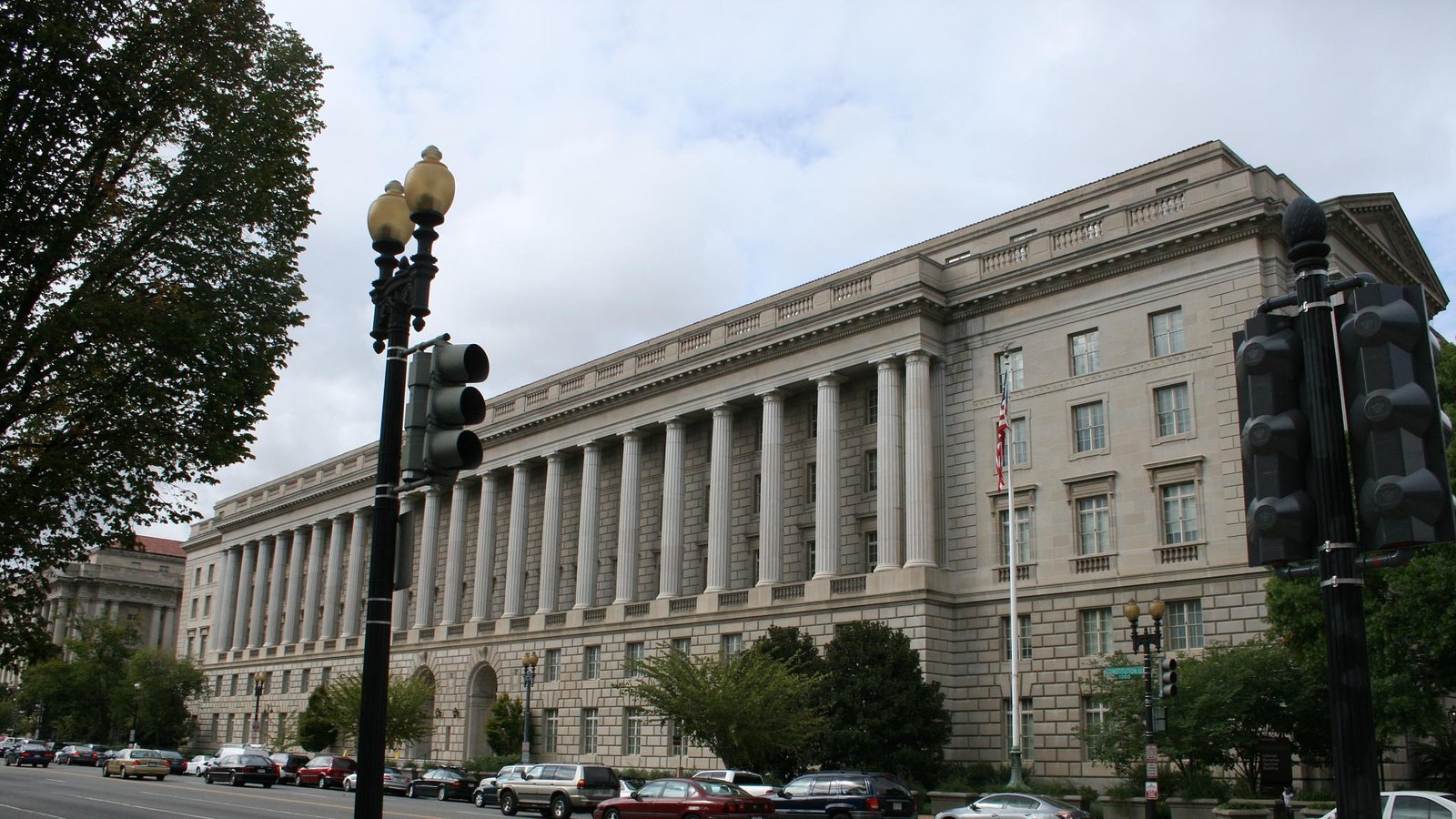 The Internal Revenue Service Building on Constitution Avenue in Washington, a long limestone colonnade running the length of the block under a white sky, with an American flag on a pole at the corner, traffic signals and a street lamp in the foreground and cars parked along the kerb.