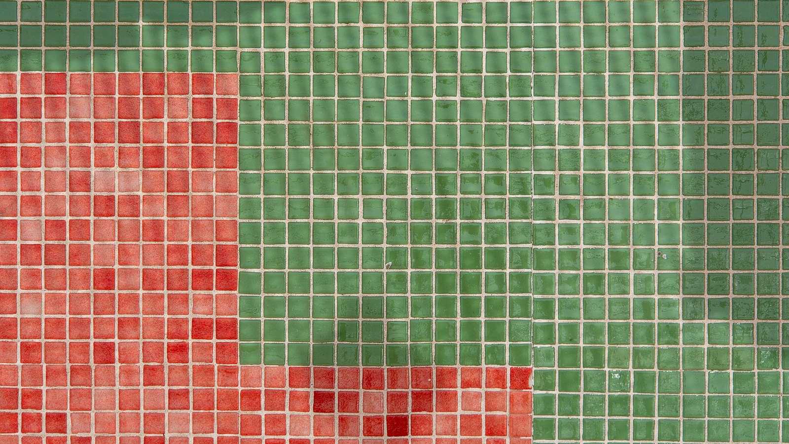 A close photograph of a wall of small square glass mosaic tiles in even rows, most of them green, with a solid block of red tiles filling the lower left and a short run of red along the bottom edge. No text, brand mark, person or place is in view.