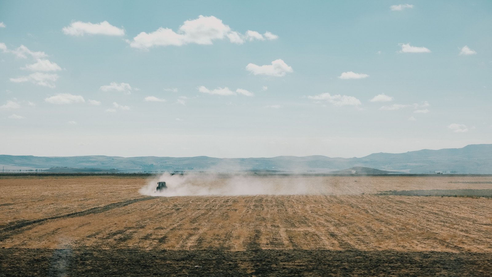 A small tractor far off across a wide dry ploughed field, trailing a long plume of dust, with low hills and a pale blue sky with scattered clouds behind.