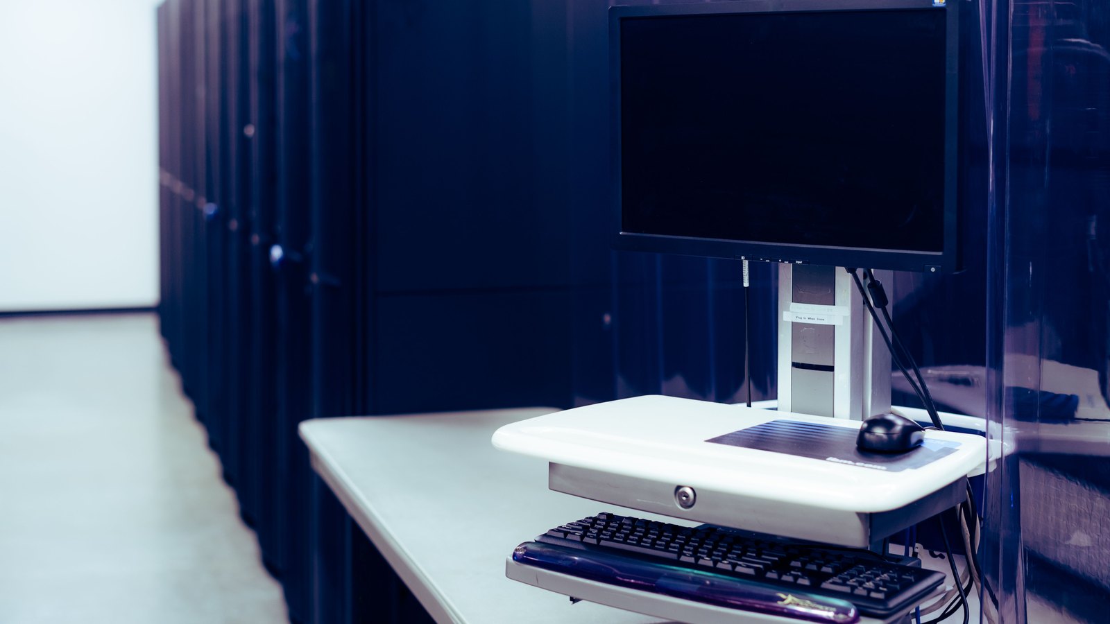 A single workstation standing at the end of a row of dark server cabinets in a data hall, a blank switched-off monitor on a white adjustable stand above a keyboard and mouse. The room is empty. No person, brand, logo or legible text appears anywhere in the frame. Stock photograph, not a photograph of anything in this story.