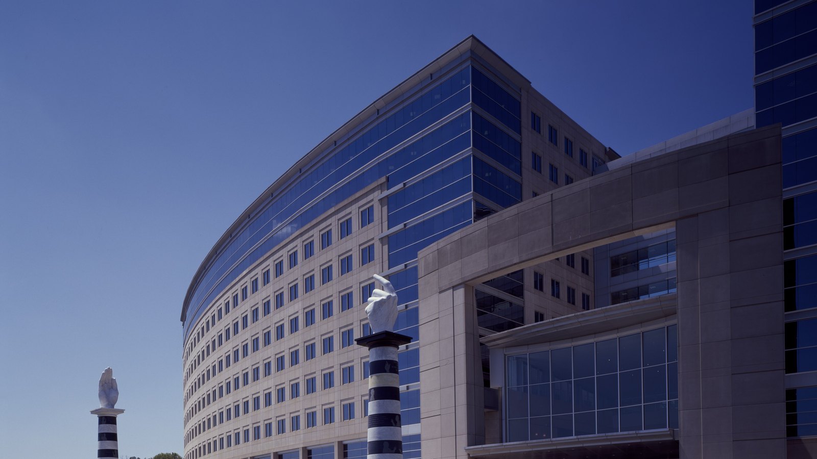 A large modern federal office building with a long curved facade of blue tinted glass and pale stone, seen from below against a clear deep blue sky, with two striped stone columns carrying white sculpted hands standing in front of it. No people, vehicles, signage or lettering are in the frame.