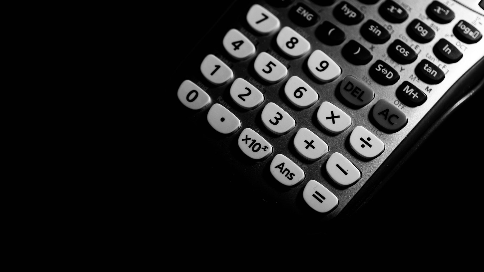 A calculator photographed at an angle against a black background, the pale number keys and the function keys lit from one side and the left half of the frame in darkness. No brand name, date or document appears anywhere in the picture.