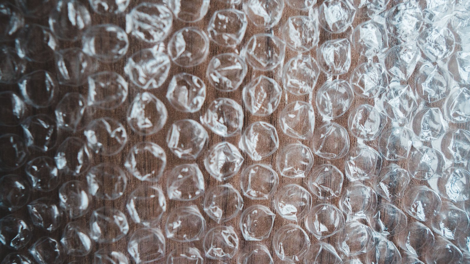 A sheet of clear bubble wrap laid flat over dark wood and filling the whole frame, its rows of round air pockets catching the light, with the brown grain of the surface showing through between them. No text, brand mark, person or place is in view.